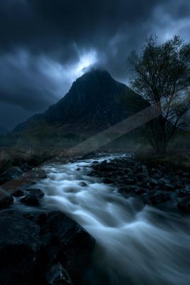 A fast-flowing river rushes over dark rocks in the foreground, leading toward a large rugged mountain partially shrouded in mist and heavy storm clouds, with a 