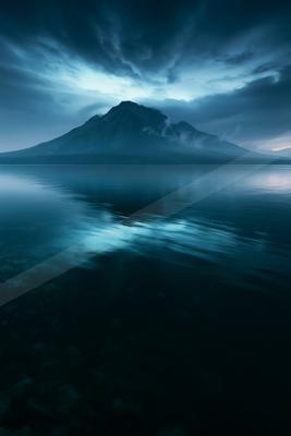 A serene lake reflects a majestic mountain under a moody twilight sky.