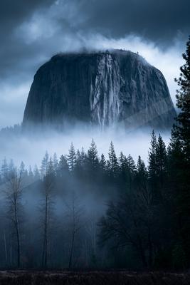 A towering mountain peak rises above a dense forest shrouded in mist, under a dark, cloudy sky.