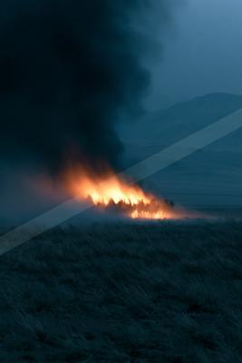 A wildfire burns intensely across a dry grassy plain at dusk, sending a massive plume of dark smoke into the blue sky with distant hills visible in the backgrou