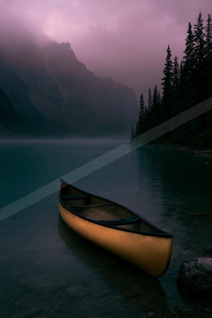 A yellow canoe rests at the edge of a calm lake with misty mountains and a pink-purple sky in the background, surrounded by tall evergreen trees.
