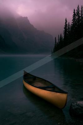 A yellow canoe rests at the edge of a calm lake with misty mountains and a pink-purple sky in the background, surrounded by tall evergreen trees.