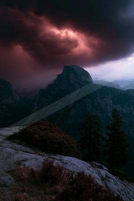 Moody atmospheric landscape with dark reddish-purple storm clouds hovering over a large granite mountain peak, with rocky foreground, shrubs and pine trees unde