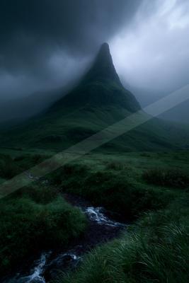 A sharp rocky mountain peak rises through thick fog and dark storm clouds, with a small stream flowing through lush green grass and vegetation in the foreground