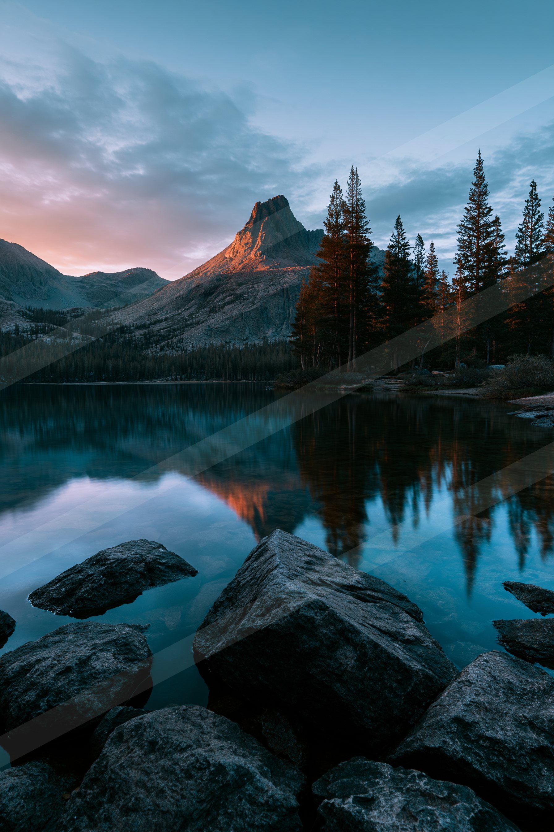 A serene lake reflects the silhouette of towering pine trees and a rugged mountain peak bathed in warm sunset light, creating a stunning mirror effect.