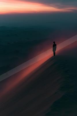 A silhouetted man stands atop a vast sand dune overlooking a sea of dunes bathed in red and orange sunset light, with atmospheric haze filling the valley below.
