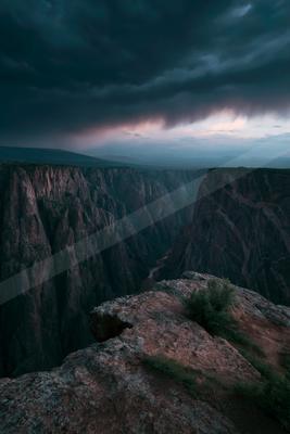 A dramatic view from a rocky cliff edge overlooking a vast, deep canyon with a winding river below, under heavy dark storm clouds with a hint of pink light on t