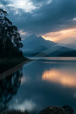 A tranquil lake reflects the vibrant hues of a sunset sky with silhouetted trees and distant mountains.