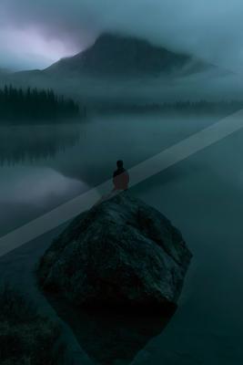 A solitary figure sits in meditation pose on a large rock surrounded by still, misty water, with a forested mountain rising into the clouds in the background.