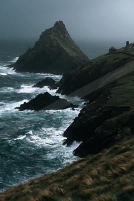 A rugged green and rocky coastal cliff juts into a turbulent sea with white-capped waves crashing against dark rocks under a foggy, overcast sky.