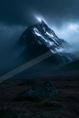 A sharp, snow-covered mountain peak rises through thick, dark clouds and fog, illuminated from above, with a rocky foreground under a moody sky.