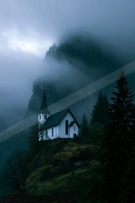 A white church with a tall steeple sits on a grassy mountain ridge surrounded by dense fog and dark evergreen trees.