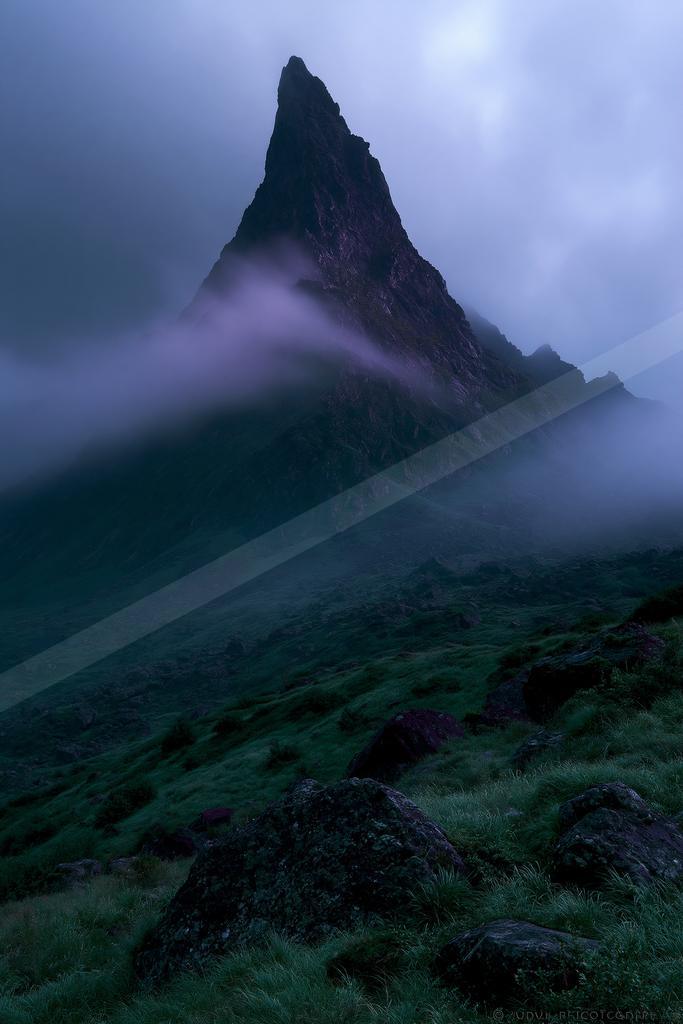 A sharp rocky mountain peak emerges through thick purple-tinged mist and low clouds, with a lush green rocky slope in the foreground under a moody overcast sky.