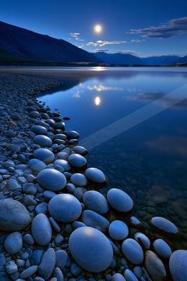 A serene night scene featuring smooth pebbles on a lakeshore, a calm reflective lake mirroring the bright moon and mountains under a deep blue sky.
