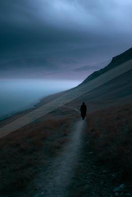A lone figure in dark clothing walks along a winding dirt path on a barren hillside, with a large flock of animals scattered below near a foggy valley under dra