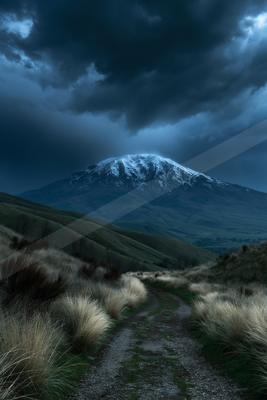 A winding dirt path leads through tall dry grass toward a large snow-capped mountain under a dark, stormy sky with dramatic clouds.