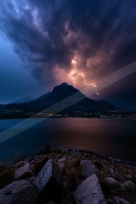 A powerful lightning bolt strikes near a dark mountain peak during a thunderstorm, illuminating dramatic purple clouds above a calm lake and rocky foreground sh