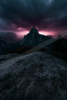 A rugged granite mountain peak rises against a dramatic sky filled with dark storm clouds illuminated by intense magenta and pink light at the horizon, with a l