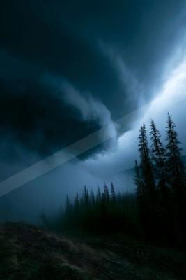 Moody atmospheric landscape with dark storm clouds swirling over a dense evergreen forest shrouded in thick fog, with a patch of bright light breaking through o