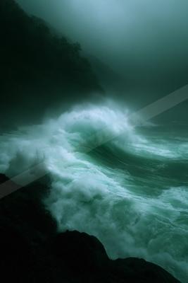 A powerful turquoise wave crashes against dark rocks in a misty, foggy cove with forested cliffs in the background.