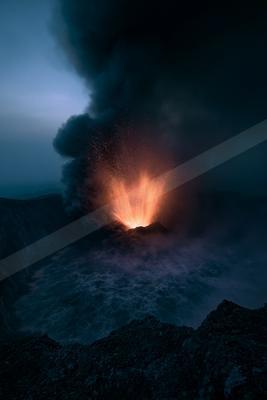 A powerful volcanic eruption at night, with bright orange lava exploding upwards and a massive dark ash plume rising into the sky over a rocky crater.