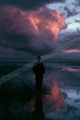 A silhouetted person stands in a wetland at dusk, gazing at massive, dramatic pink and purple storm clouds reflected in the water below.