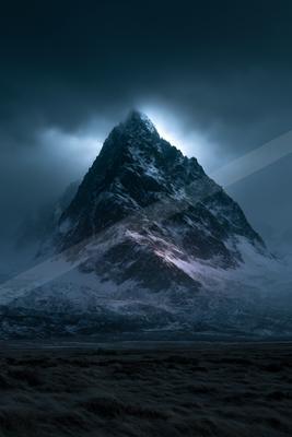 A towering, jagged mountain covered in snow rises dramatically against a dark, cloudy sky, with light breaking through the clouds near the summit, above a dark 