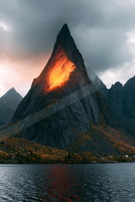A sharp rocky mountain peak catches the last warm light of sunset, glowing orange against dark storm clouds, with its reflection in the calm water of a fjord be