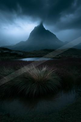 A dark, moody landscape featuring a towering mountain peak silhouetted against stormy skies, reflected in a calm lake surrounded by grasses.