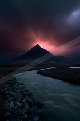 A dark rocky mountain peak rises against a dramatic red and purple stormy sky, with a winding river flowing through a barren foreground landscape.