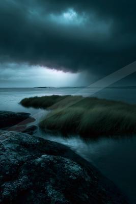 Moody coastal landscape featuring dark storm clouds, a patch of tall grass on a small islet, calm water, and large wet rocks in the foreground under a dramatic 