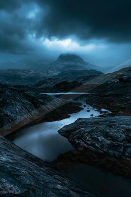 A serene twilight scene featuring a calm lake surrounded by rugged terrain, with a distant mountain peak partially shrouded in misty clouds.
