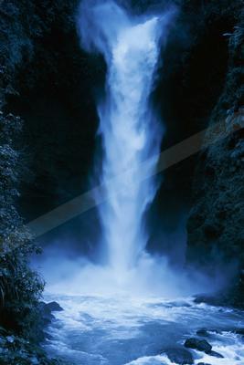 A powerful waterfall with bright blue-white water cascades down a dark rocky cliff surrounded by dense green foliage, creating mist at the base where turbulent 