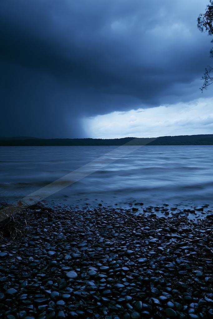A dramatic stormy scene over a large lake with dark heavy clouds, visible rain in the distance, a pebble-covered shore in the foreground, and a distant forested
