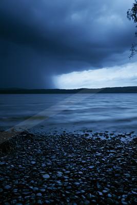 A dramatic stormy scene over a large lake with dark heavy clouds, visible rain in the distance, a pebble-covered shore in the foreground, and a distant forested
