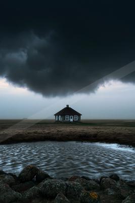 A lone cottage stands in an open field under ominous storm clouds, with rippling water and rocky foreground.