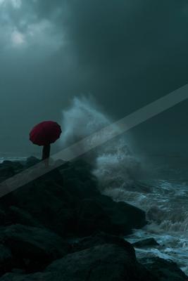 A solitary figure holding a bright red umbrella stands on dark rocks as a powerful white wave crashes dramatically against the shore under a dark stormy sky.