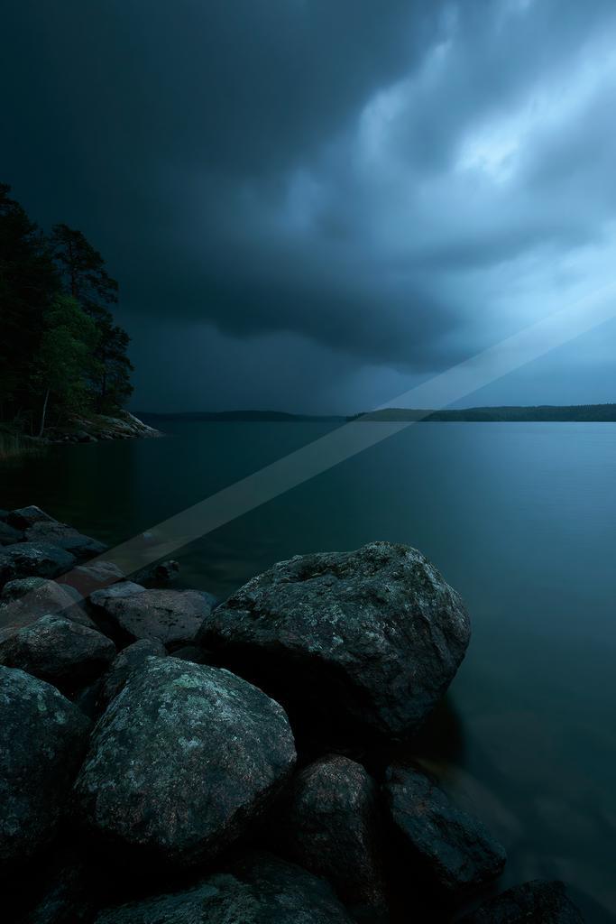 Large dark rocks in the foreground on the shore of a calm lake under a sky filled with heavy storm clouds, with a forested landmass visible on the left.