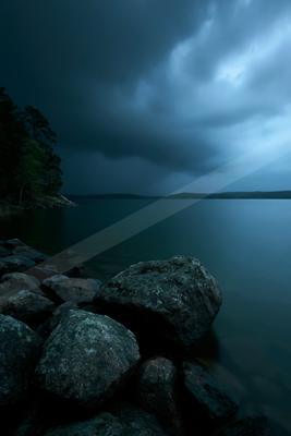 Large dark rocks in the foreground on the shore of a calm lake under a sky filled with heavy storm clouds, with a forested landmass visible on the left.