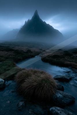 A dramatic, fog-shrouded mountain peak rises above a flowing stream in a rugged, grassy landscape under a moody blue sky.