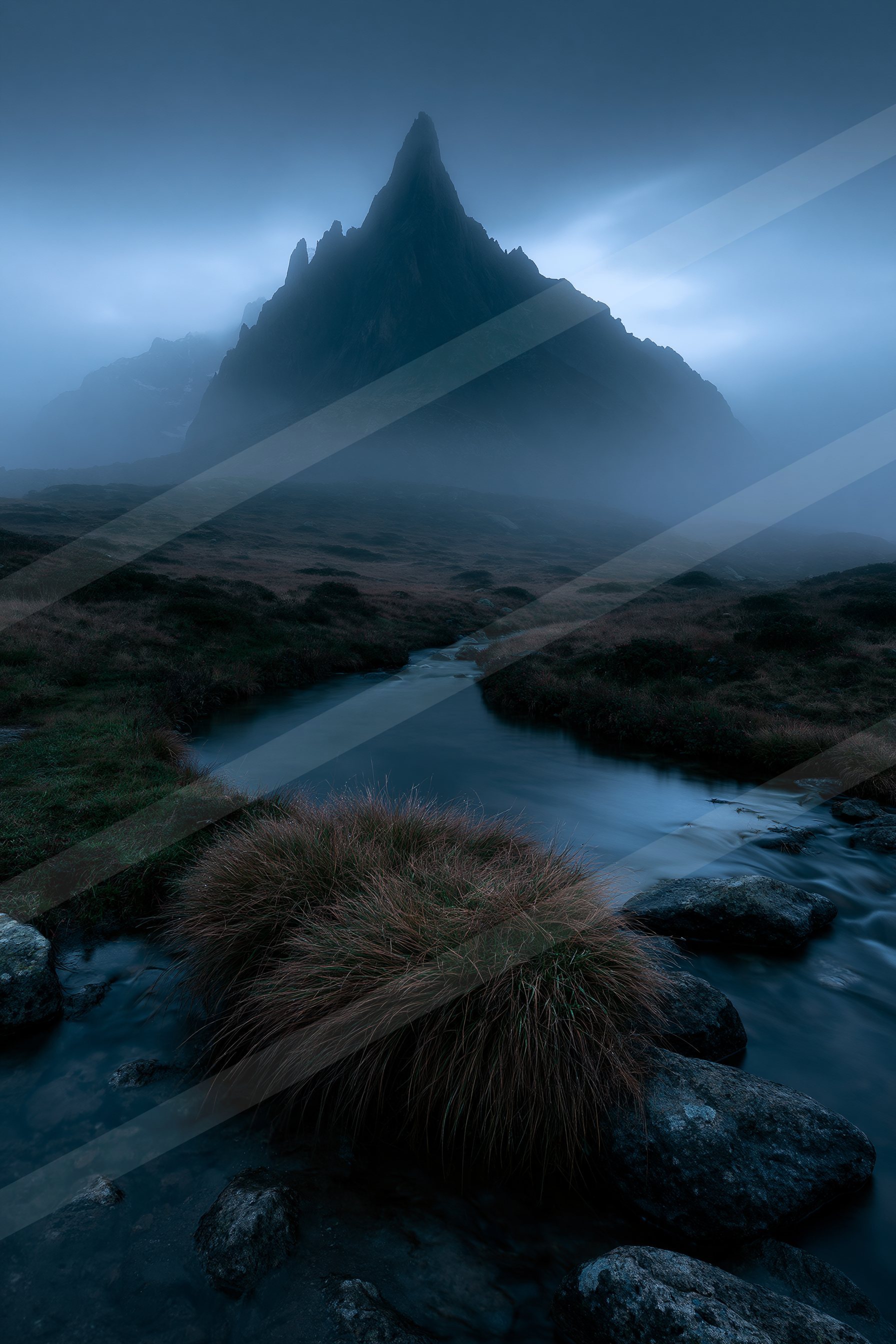 A towering mountain peak shrouded in mist overlooks a serene stream winding through rugged terrain.
