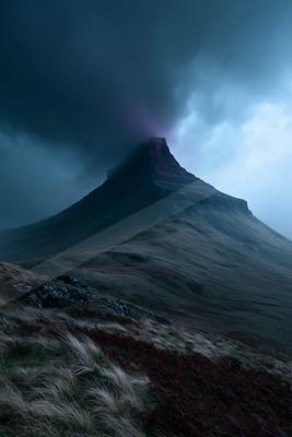 A steep rocky mountain peak rises into dark, heavy storm clouds with a hint of purple light at the summit, while the foreground shows a grassy hillside with roc