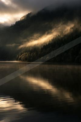 A serene lake reflects the golden glow of sunlight filtering through misty clouds on a forested mountain slope.