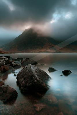 A large rocky mountain partially shrouded in mist rises above a calm lake, with large dark rocks in the foreground under a dramatic cloudy sky.