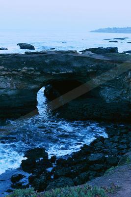 A large natural rock arch spans over turbulent blue ocean waves and dark rocks on a rugged coastline under a pale sky.