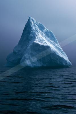 A towering white and blue iceberg rises from the dark, rippled waters of the ocean under a foggy, overcast sky.