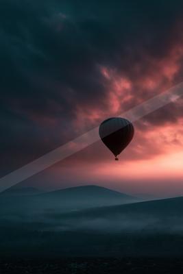 A dark hot air balloon floats in a dramatic sky filled with pink, red, and dark blue clouds above a sea of fog covering rolling hills at sunset.