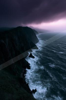 Aerial view of a rugged dark cliff coastline meeting a turbulent ocean with white foam waves under a moody purple and dark cloudy sky at dusk.