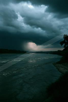 Moody landscape with dark storm clouds over a wide flowing river, light breaking through the clouds in the distance, with a tree-lined shore on the right.