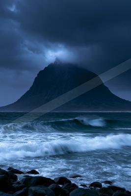 Powerful ocean waves with white foam crash towards a rocky foreground under a dark, cloudy sky, with a large rocky mountain partially shrouded in mist in the ba